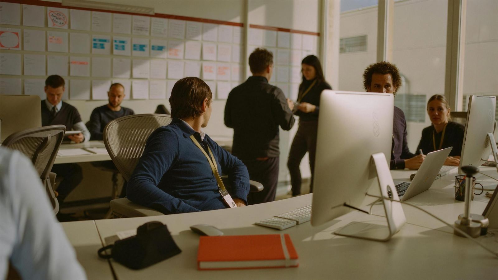 An embedded recruiter with a visitor lanyard working among an in-house HR team, a wall of role cards behind