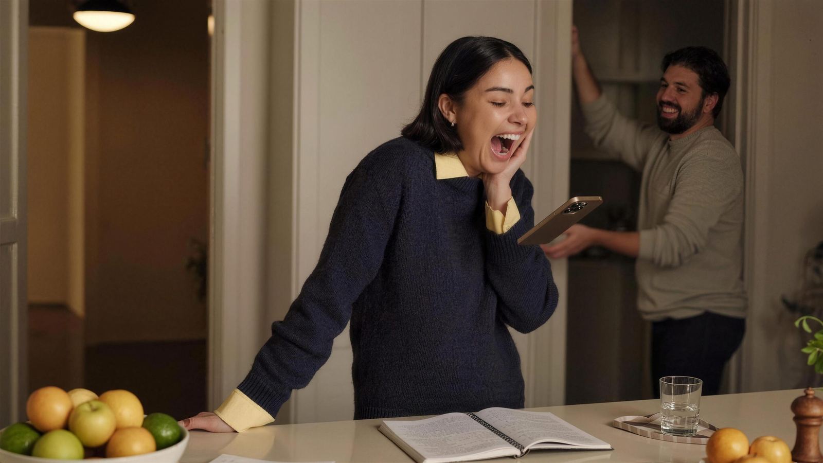 A woman laughing at her phone in a warm kitchen as her partner comes through the doorway